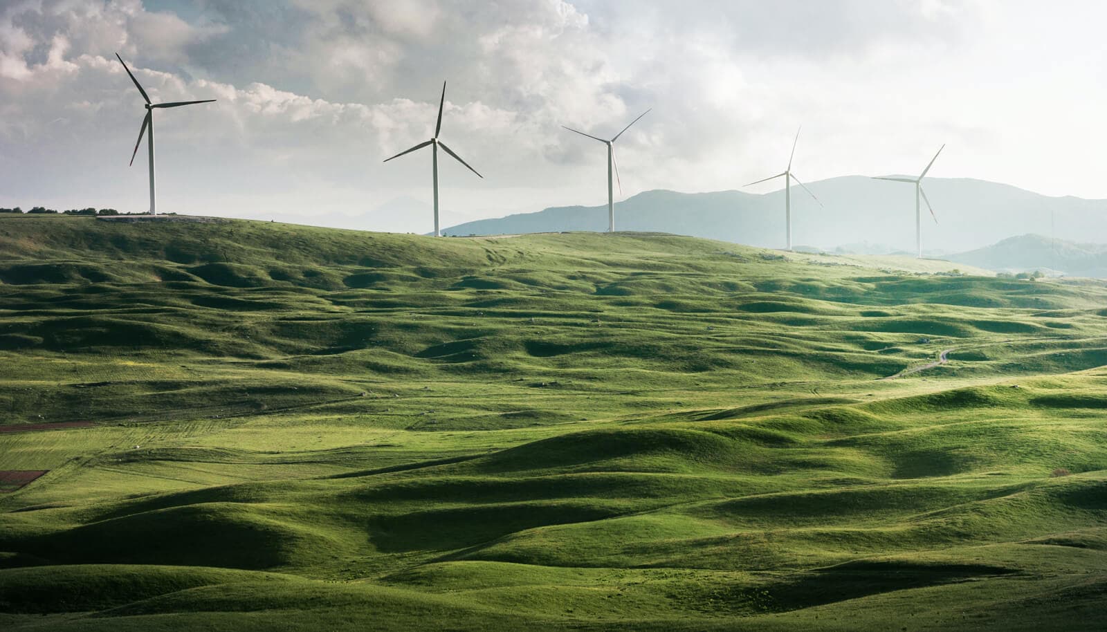 A wind farm with 6 windmills, rolling hills, and a cloudy gray sky in the background.