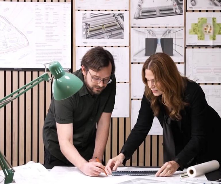 people leaning over a desk in front of a wall of architecture renderings