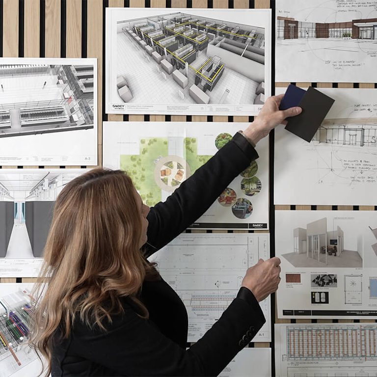 person holding paint swatches up to a wall of architecture drafts and renderings