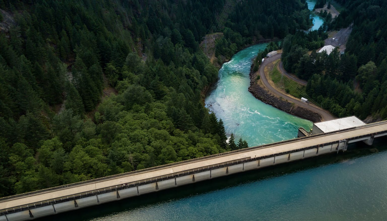 A drone shot of a dam from the high side with a river and trees in the background.