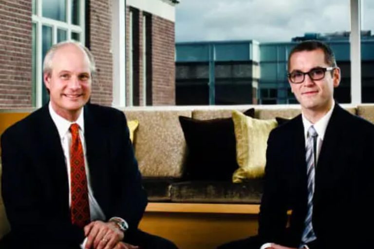 Two men sitting in front of an office window wearing suits.