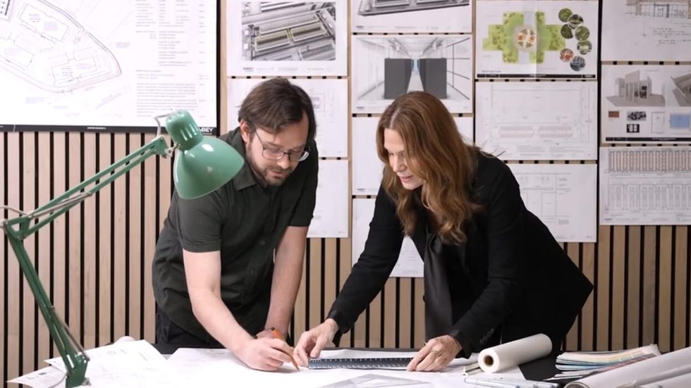 Two people leaning over a desk with papers sprawled across it and papers pinned on the wall in the background.