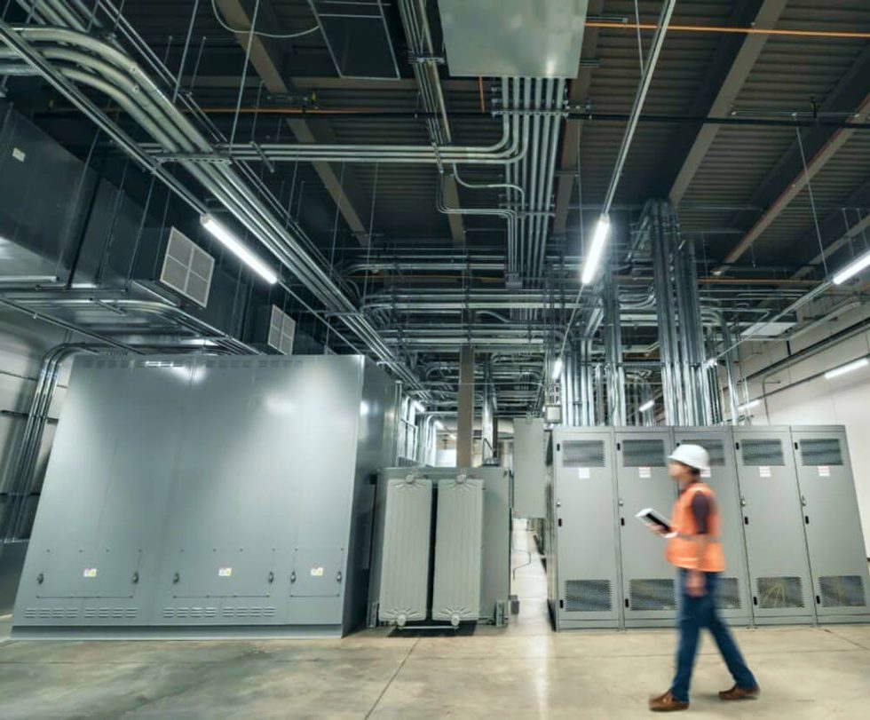 A person wearing a vest and hard hat walking through an industrial space with servers behind them.
